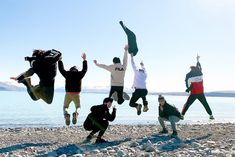 a group of people jumping up into the air on top of a sandy beach next to water