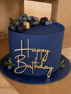 a blue birthday cake with berries and the words happy birthday written on it in gold lettering