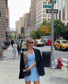 a woman in a blue dress and black blazer is walking down the street with a cup of coffee