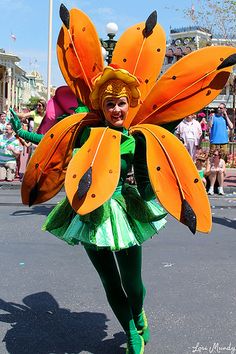 a woman in a green and orange costume is walking down the street with large wings on her head