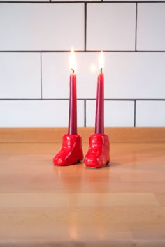 two candles with red shoes on them sitting on a wooden table in front of a white brick wall