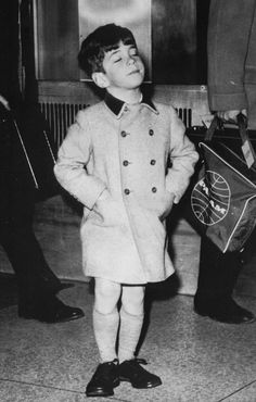 a young boy in a coat and hat standing on the sidewalk with his hands behind his back