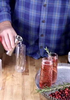 a person pouring some wine into two glasses on a table with cranberries and rosemary