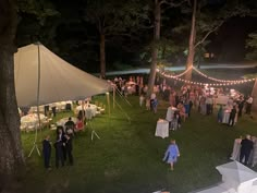 a large group of people standing around tables and chairs in the grass at an outdoor event