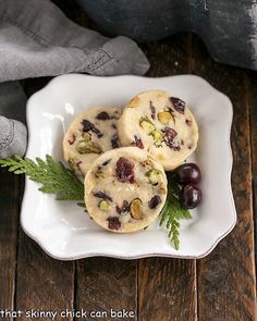 three cookies on a white plate with green leaves and berries in the middle, sitting on a wooden table