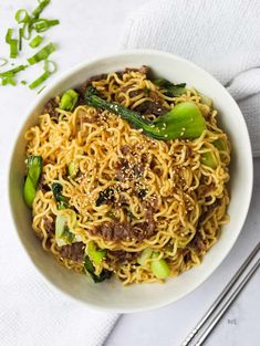 a white bowl filled with noodles and veggies on top of a table next to chopsticks