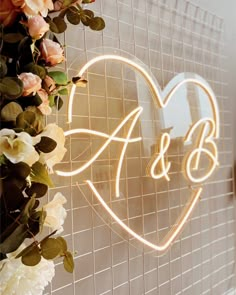 a heart shaped neon sign with flowers and greenery on the wall next to it