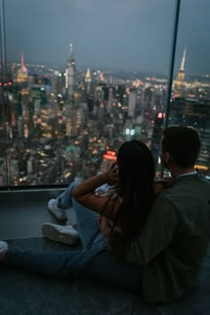 a man and woman sitting on top of a tall building looking at the city lights