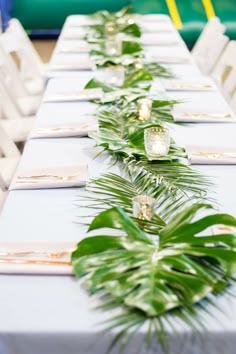 the long table is set with white chairs and green plants on it's sides