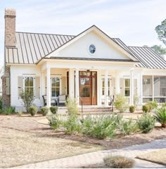 a white house with a metal roof and front porch is surrounded by greenery, shrubs and trees