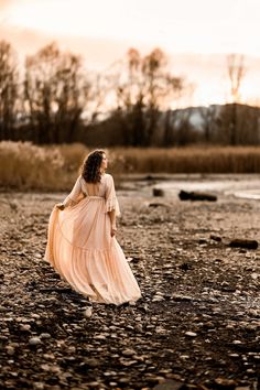 a woman in a long dress walking across a field next to a body of water