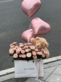 a teddy bear sitting on top of a box filled with pink roses and heart shaped balloons