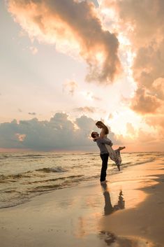 a man holding a woman on top of a beach next to the ocean at sunset