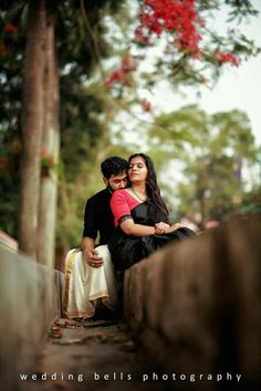 a man and woman sitting next to each other on a wooden bench in front of trees