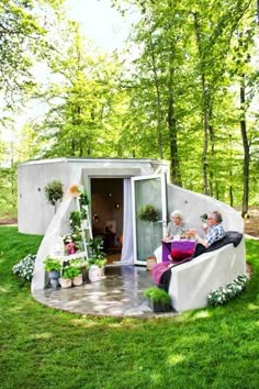 two people are sitting on the outside of a small house in the grass with potted plants