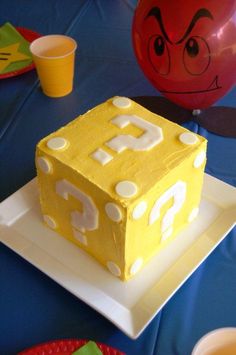 a cake that looks like a pacman block on a plate with balloons in the background