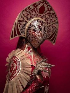 a woman in a red and gold costume with a fan on her head, against a pink background