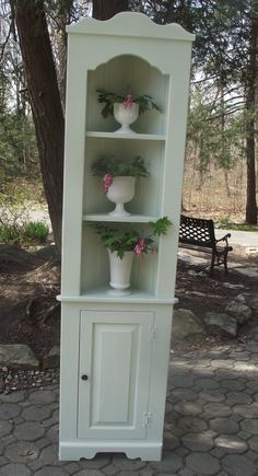 a white china cabinet with flowers on top and potted plants in the bottom shelf