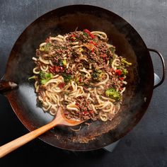 a wok filled with noodles and meat on top of a black table next to a wooden spoon