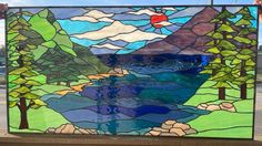 a hand made stained glass window with trees and water in the background, on a wooden bench