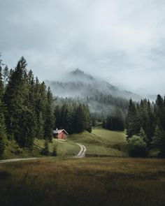 a small house in the middle of a field with trees on both sides and fog hanging over the mountains behind it