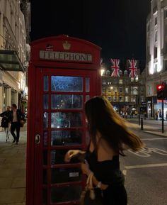 a woman is standing in front of a red phone booth on the street at night