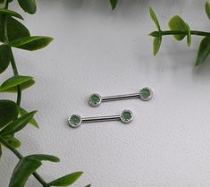 two surgical steel barbells with green crystal stones on each one, surrounded by leaves