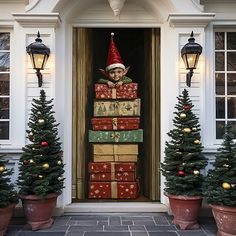 an elf is sitting on top of christmas presents in front of a house with potted trees