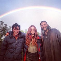 three people standing in front of a rainbow with one person wearing a black coat and the other holding a red jacket