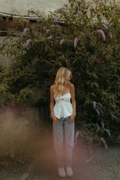 a woman standing in front of a bush with purple flowers on the bushes behind her