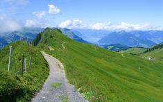 a dirt road on the side of a grassy hill with mountains in the background and clouds in the sky