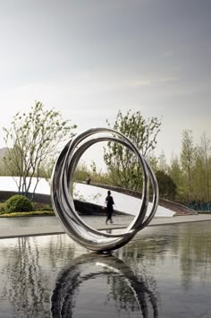 a person walking past a sculpture in the middle of a park with water reflecting off it's surface