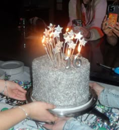 two people are reaching for a birthday cake with candles on it and one person is holding a cell phone