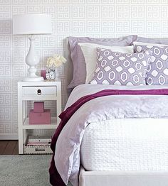 a white bed topped with purple pillows next to a night stand and nightstand filled with flowers