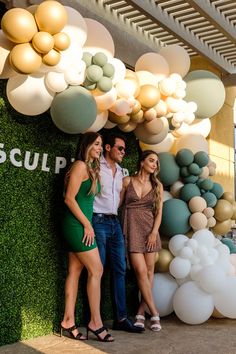 two women and a man standing in front of a wall with balloon decorations on it