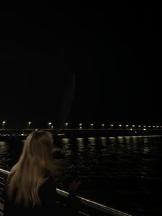 a woman is sitting on a bench looking at the water and lights in the distance