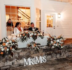 a bride and groom are sitting on a bench with flowers in front of the sign