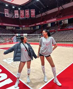 two women standing on a basketball court holding hands and talking to each other, with the floor covered in red bleachers