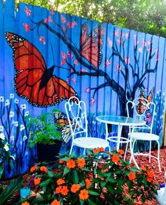 a blue fence with orange flowers and two white chairs in front of the painted wall