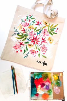 a bag with flowers painted on it next to some paintbrushes and watercolors