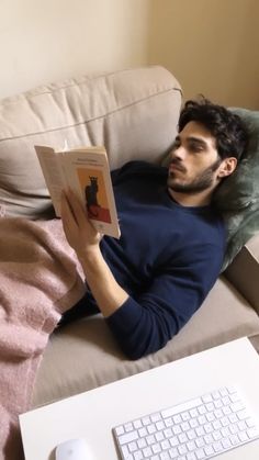 a man laying on a couch reading a book next to a keyboard and computer mouse