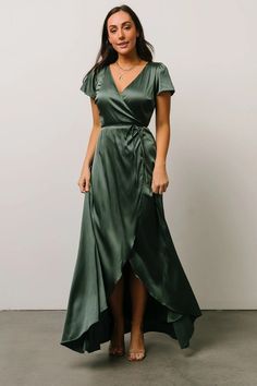 a woman wearing a green wrap dress with short sleeves and side slits, standing in front of a white wall