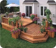 a wooden deck with potted plants and flowers on the bottom level, in front of a house