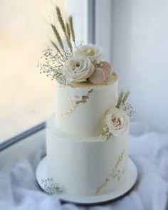 a three tiered white cake with flowers and feathers on the top is sitting in front of a window