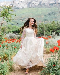 a woman in a white dress is walking through the field with red flowers and mountains behind her