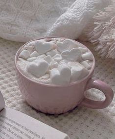 a pink cup filled with marshmallows on top of a bed next to a book