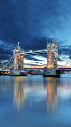 the tower bridge is lit up at night with lights reflecting in the water and clouds