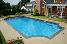 an empty swimming pool in front of a house with red flowers on the lawn and patio furniture