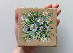 a hand holding up a small piece of art with flowers painted on it's side