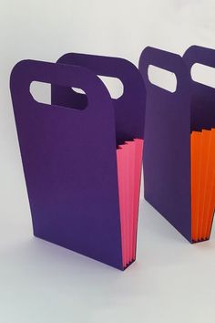 two purple and one orange paper bags on a white surface with the handles folded down